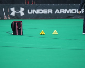 A sports field features a black and pink sports bag, two pyramids of yellow balls, and a portion of a person in dark athletic gear. The background displays a banner with the Under Armour logo.