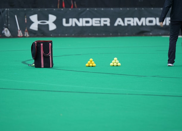 A sports field features a black and pink sports bag, two pyramids of yellow balls, and a portion of a person in dark athletic gear. The background displays a banner with the Under Armour logo.
