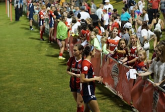 The image features a lively gathering of young soccer players and fans next to a field. Several female athletes in red and blue uniforms interact with an enthusiastic crowd standing behind a barrier. Some people in the crowd are holding objects, like pens and paper, possibly for autographs.