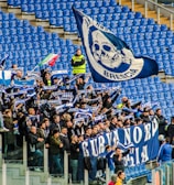 A group of enthusiastic sports fans are gathered in a stadium, holding flags, banners, and scarves that display blue and white colors and symbols. The stands are mostly empty except for this section, and the fans appear to be cheering and waving their items energetically.