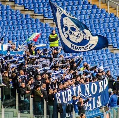 A group of enthusiastic sports fans are gathered in a stadium, holding flags, banners, and scarves that display blue and white colors and symbols. The stands are mostly empty except for this section, and the fans appear to be cheering and waving their items energetically.