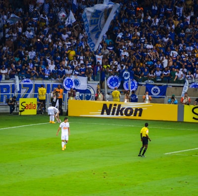 A crowded stadium with enthusiastic fans cheering, many of whom are wearing blue jerseys and holding flags. On the field, a referee in a yellow and black uniform is near the sideline, observing the match. Advertising boards for various brands like Nikon and Sadia line the edge of the field, while a player in white uniform walks nearby.