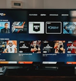 A television screen displaying the ESPN+ streaming interface with various sports-related shows and categories. The screen includes sections such as 'Browse by Shows' and 'Best of Drew Brees' with thumbnails of sports programs featuring athletes in action and analysts.