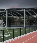 An outdoor sports facility featuring a running track and a soccer goal. The track is red, surrounded by a black metal fence, and the field is covered with artificial green turf. In the background, a basketball court is visible under a covered roof with metal supports. The sky is overcast, adding a moody atmosphere.